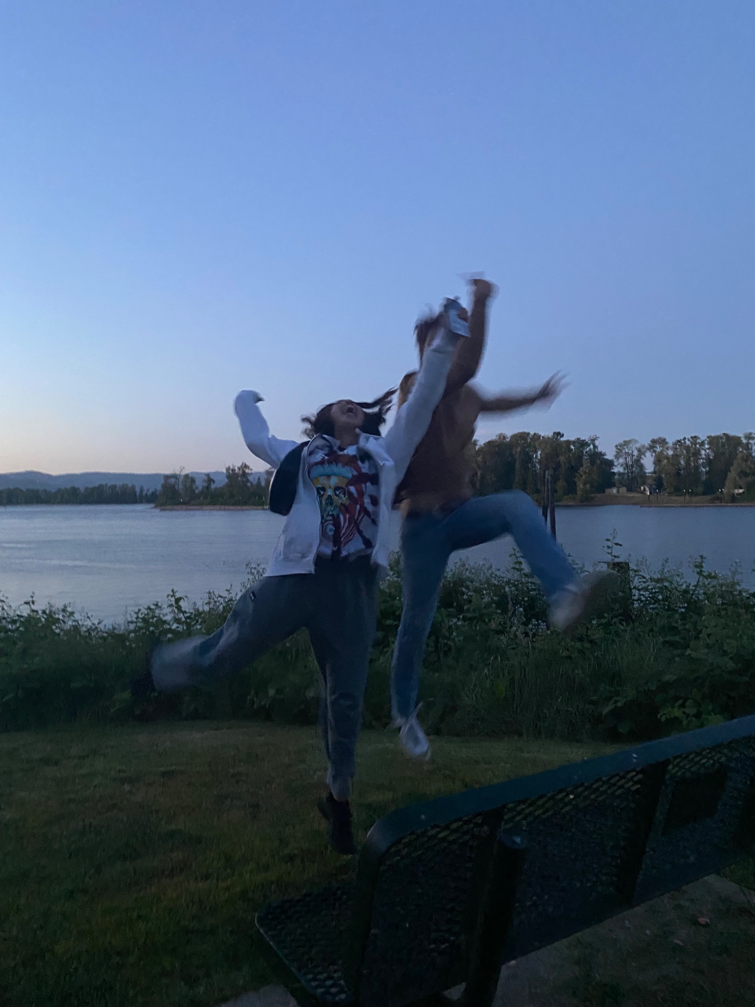 Two friends jumping joyfully beside the water at dusk